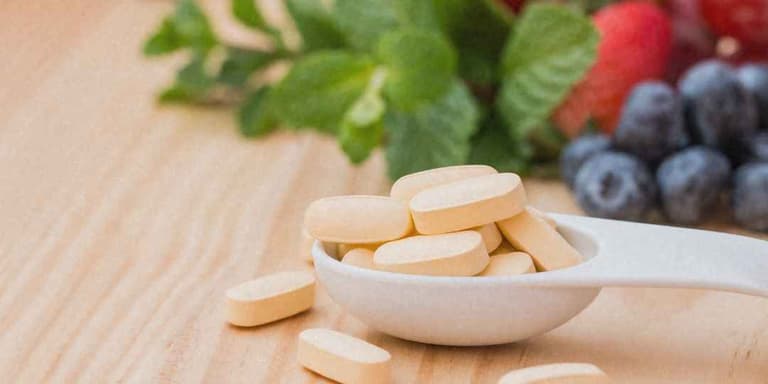 Helle Tabletten auf einem weißen Löffel, im Hintergrund Minze, Erdbeeren und Heidelbeeren.