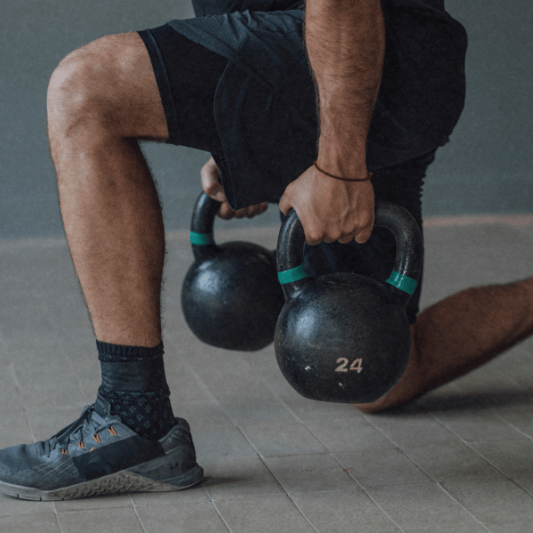 Person im Ausfallschritt hält zwei Kettlebells, Nahaufnahme von Beinen und Händen.